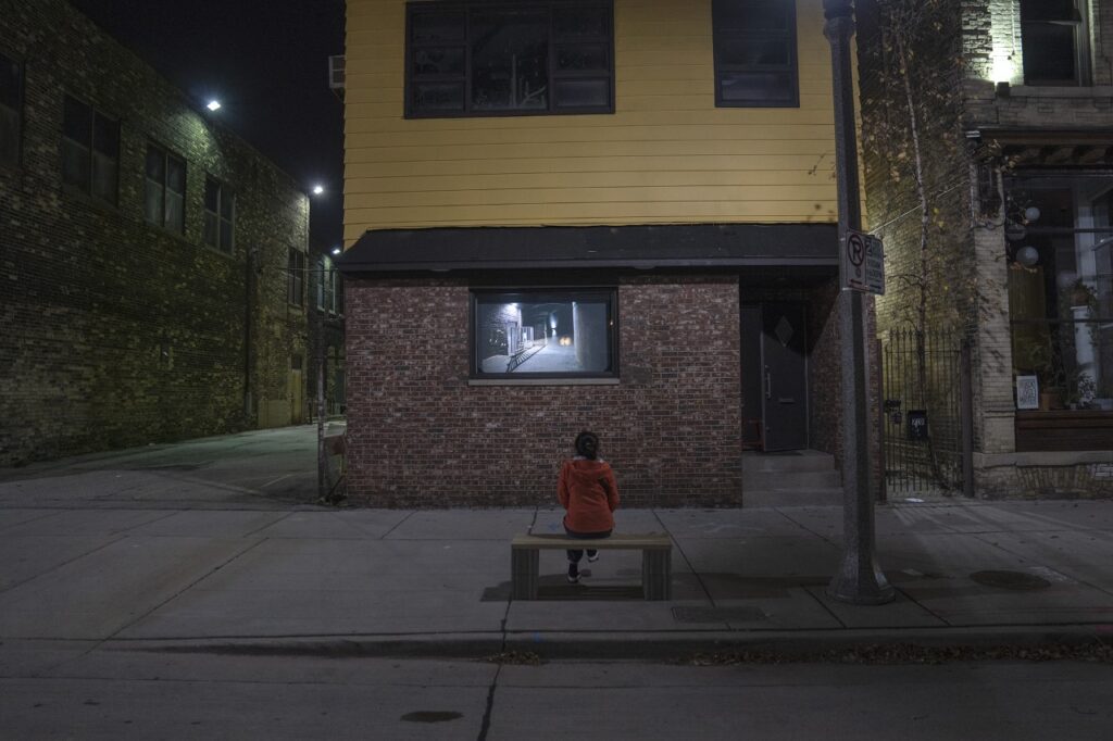 Woman sitting on bench outside The Suburban gallery, viewing Blau's artwork projected in the window.